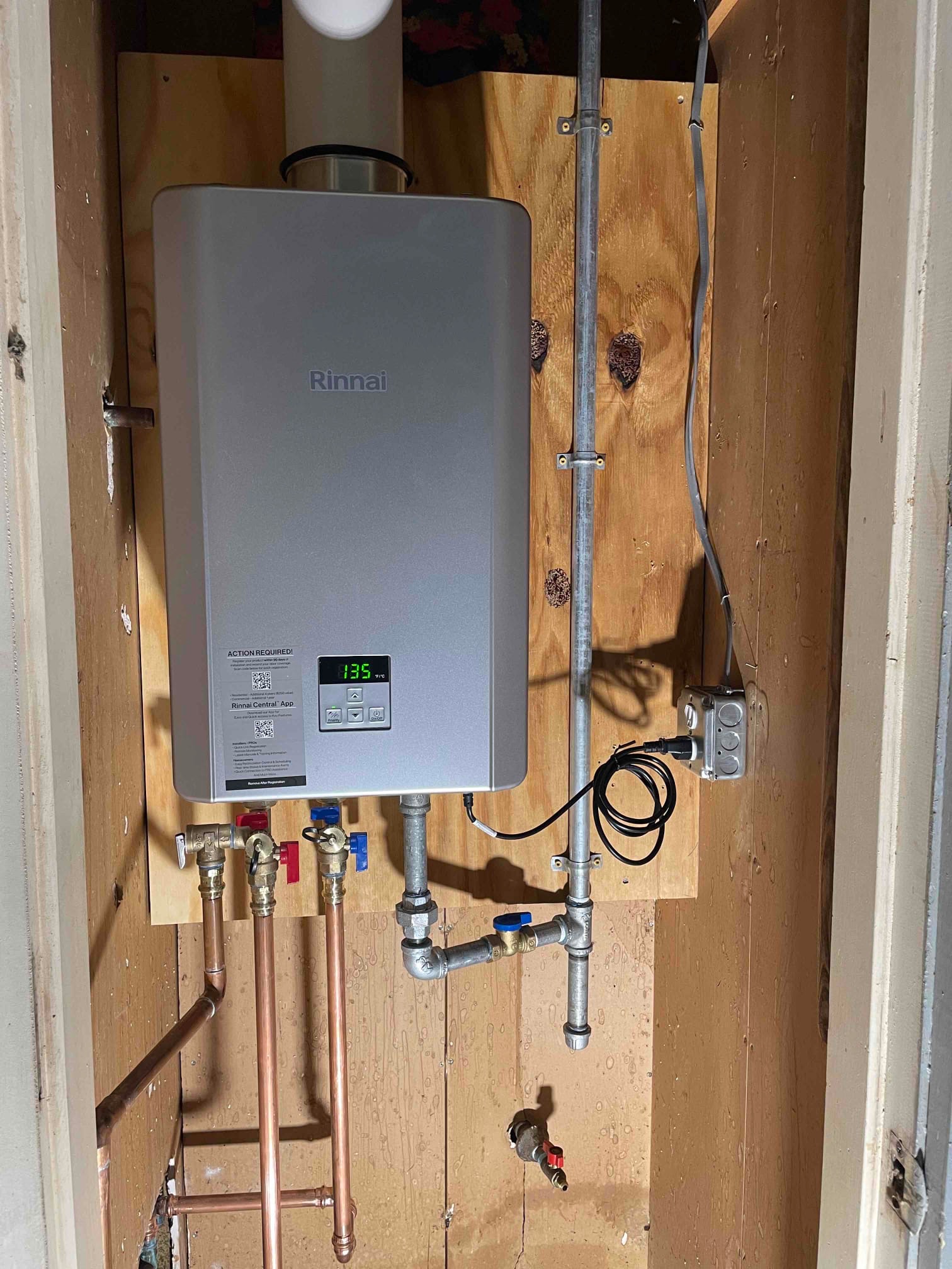 Rinnai tankless water heater mounted on a wood panel with visible plumbing connections.